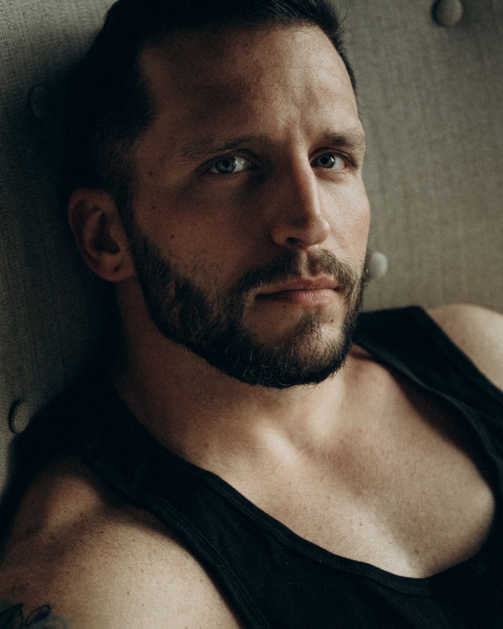 Intimate close-up portrait of a bearded man in a black tank top against a textured wall.
