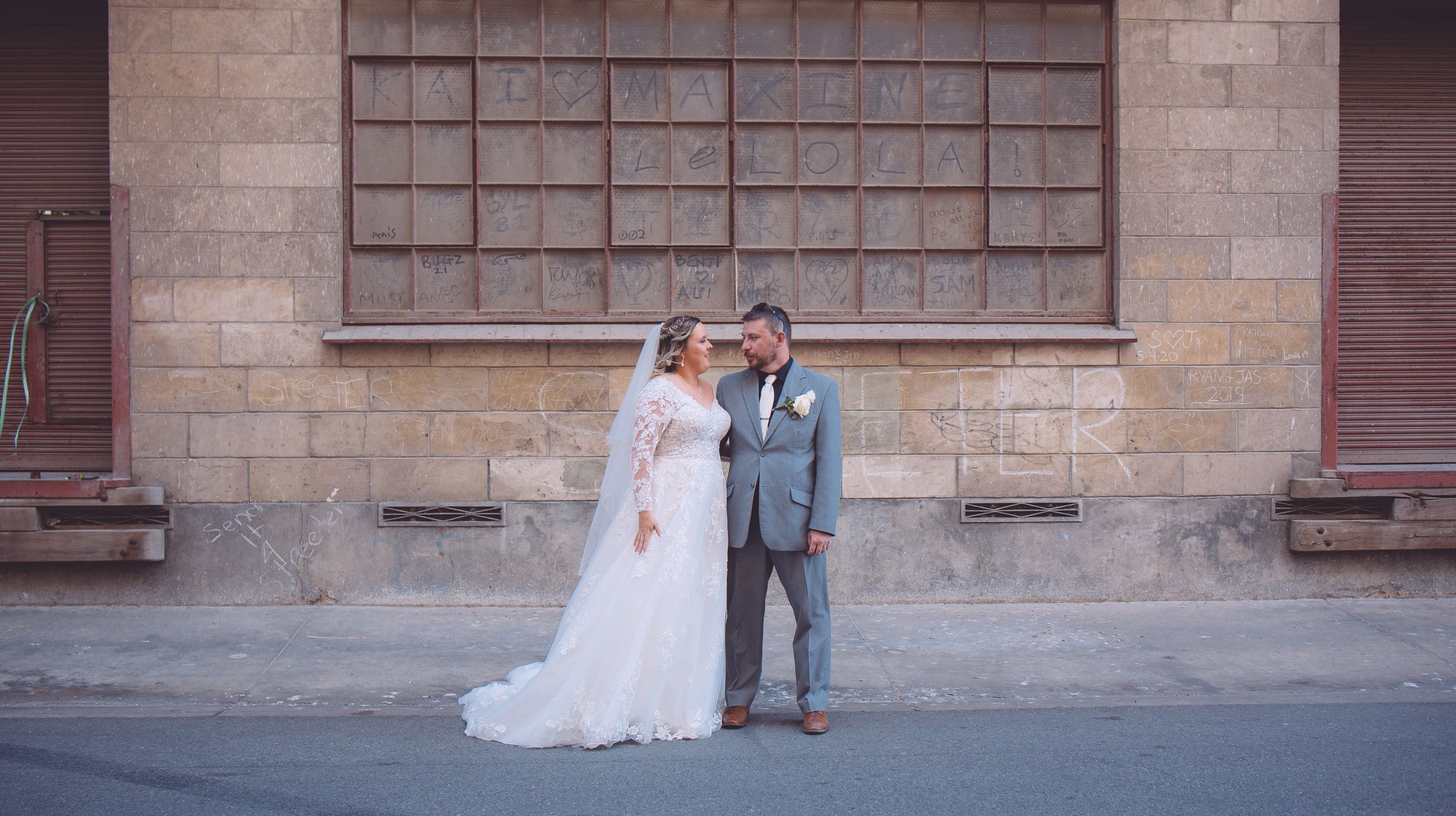 A romantic wedding couple poses against a rustic brick wall with a large industrial window.