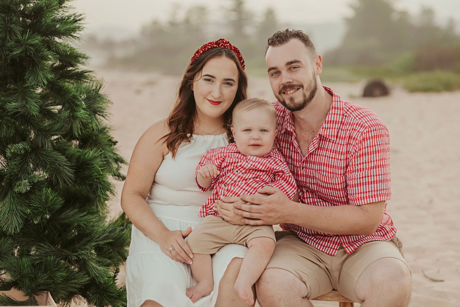 Young family Christmas beach portrait with parents and baby sitting near a decorated Christmas tree.