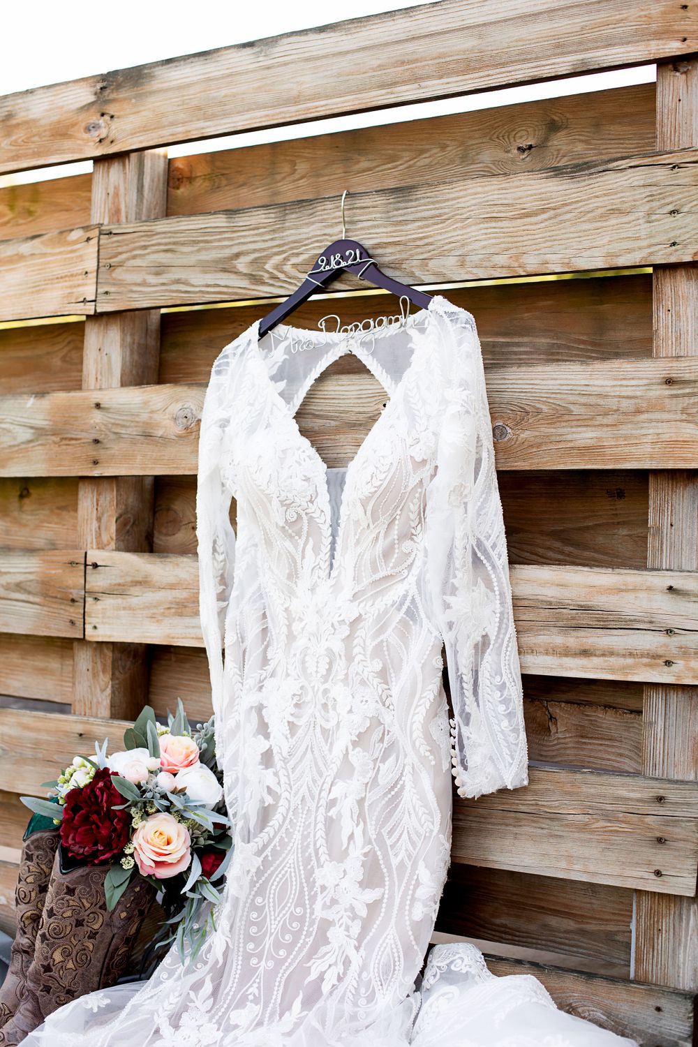 A white lace wedding dress with long sleeves hanging on wooden pallets with a floral bouquet.