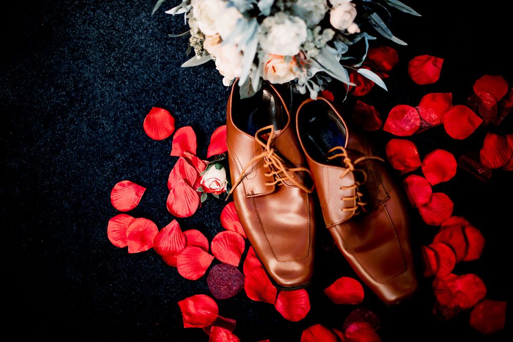 Brown leather dress shoes with wedding florals against a dark background with red bokeh lights.