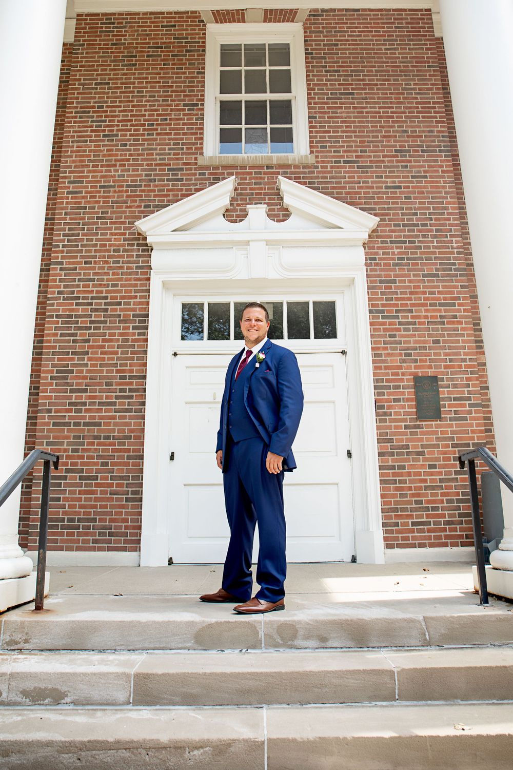 A formal navy suit pose in front of classic brick building with white double doors.