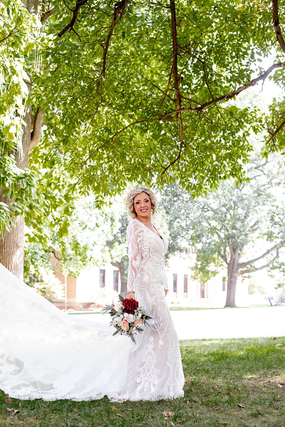 An elegant bridal portrait under green trees with lace dress and burgundy bouquet.