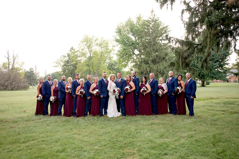 A wedding party in navy and burgundy poses on a grassy lawn during golden hour.