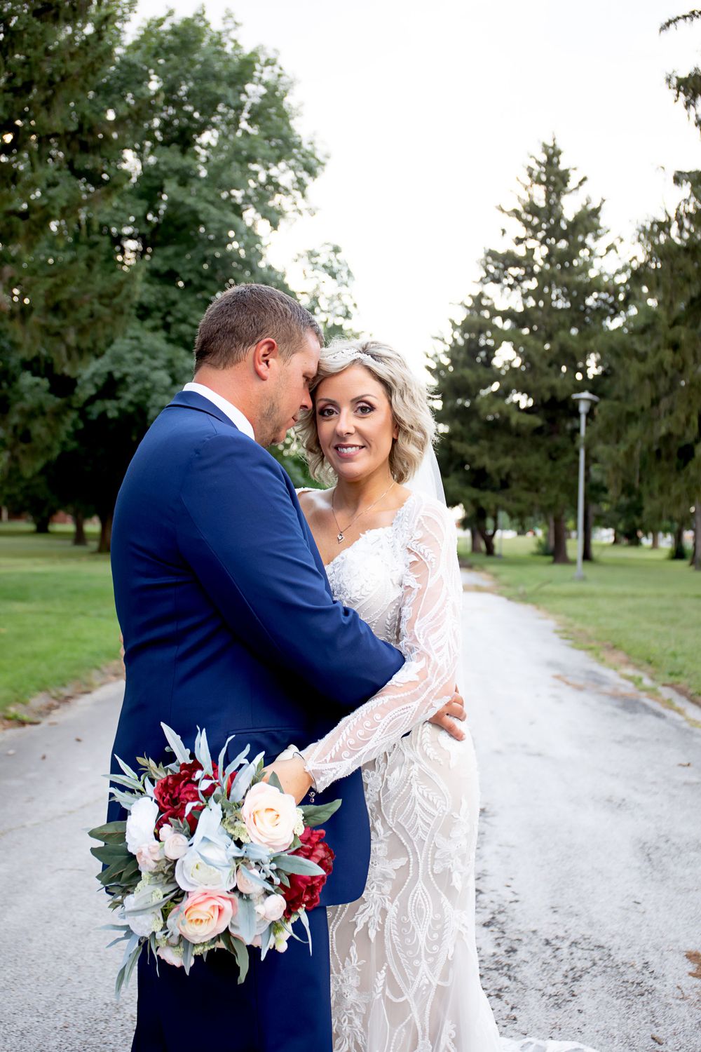 Newlyweds share intimate moment on garden path while holding elegant burgundy and cream floral bouquet.