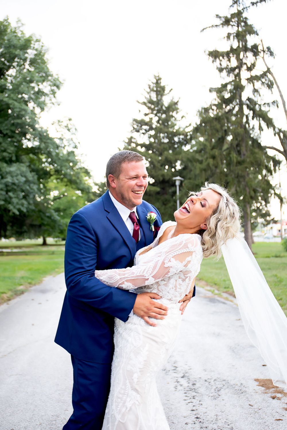 Joyful wedding couple laughing and embracing on outdoor path surrounded by evergreen trees.