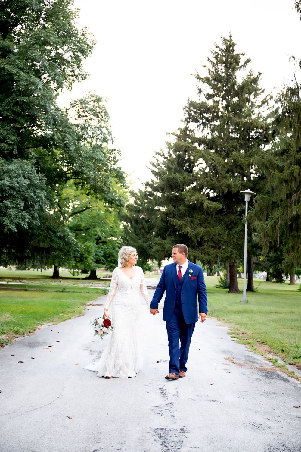 Bride and groom walking hand-in-hand down tree-lined pathway in elegant wedding attire.