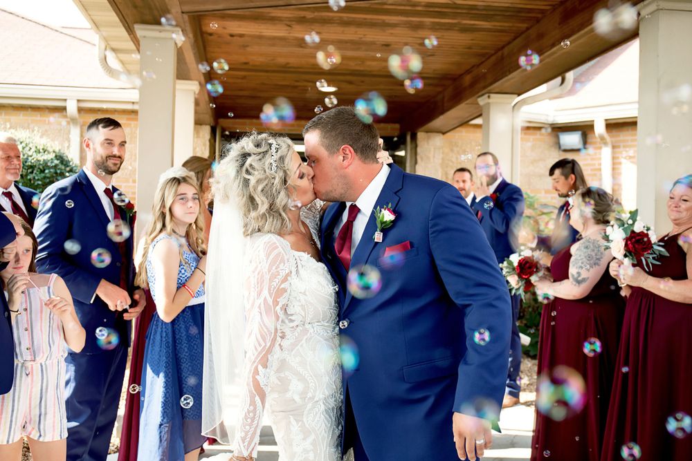 Wedding couple kissing under wooden pergola while guests shower them with bubbles.