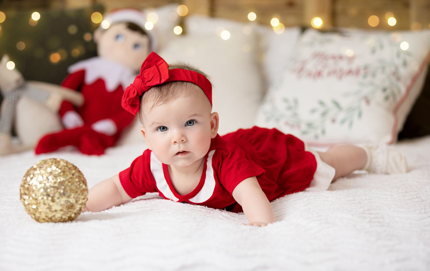 Baby in red Christmas outfit lying on white blanket next to gold ornament.