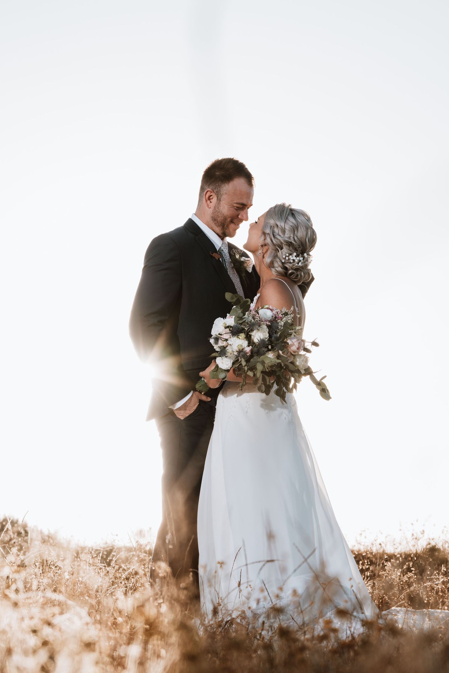 Bride and groom sharing a romantic moment in golden field, backlit by sunlight, bride holding a wildflower bouquet.