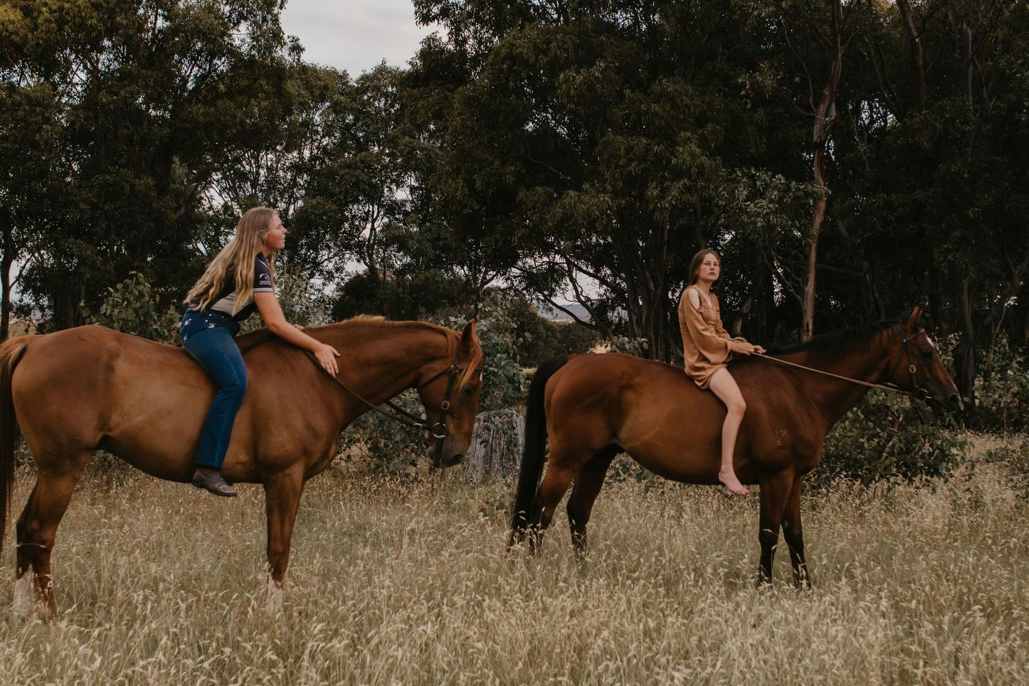 two girls riding bareback on horses through tress in a paddock on a cloudy day.