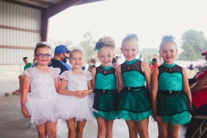 Young dancers in white and green performance costumes posing together under a covered area.