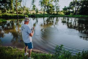 A person fishing at a tranquil pond surrounded by trees on a sunny day.