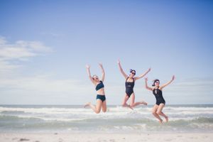 Group of friends jumping joyfully on a sunny beach with ocean waves in the background.