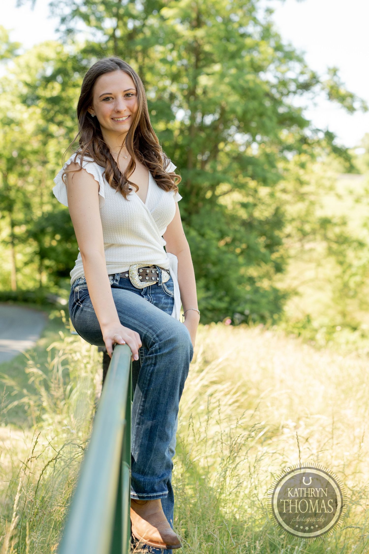 A casual outdoor portrait sitting on a green railing wearing jeans and white top.