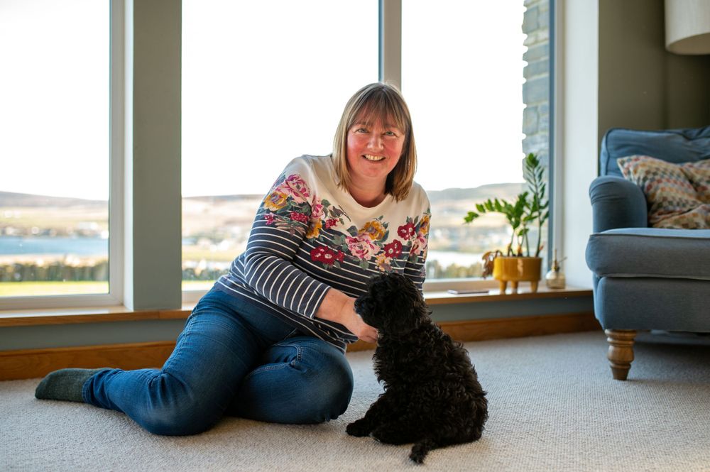 Someone in a festive sweater sits on carpet playing with a small black puppy near a window.