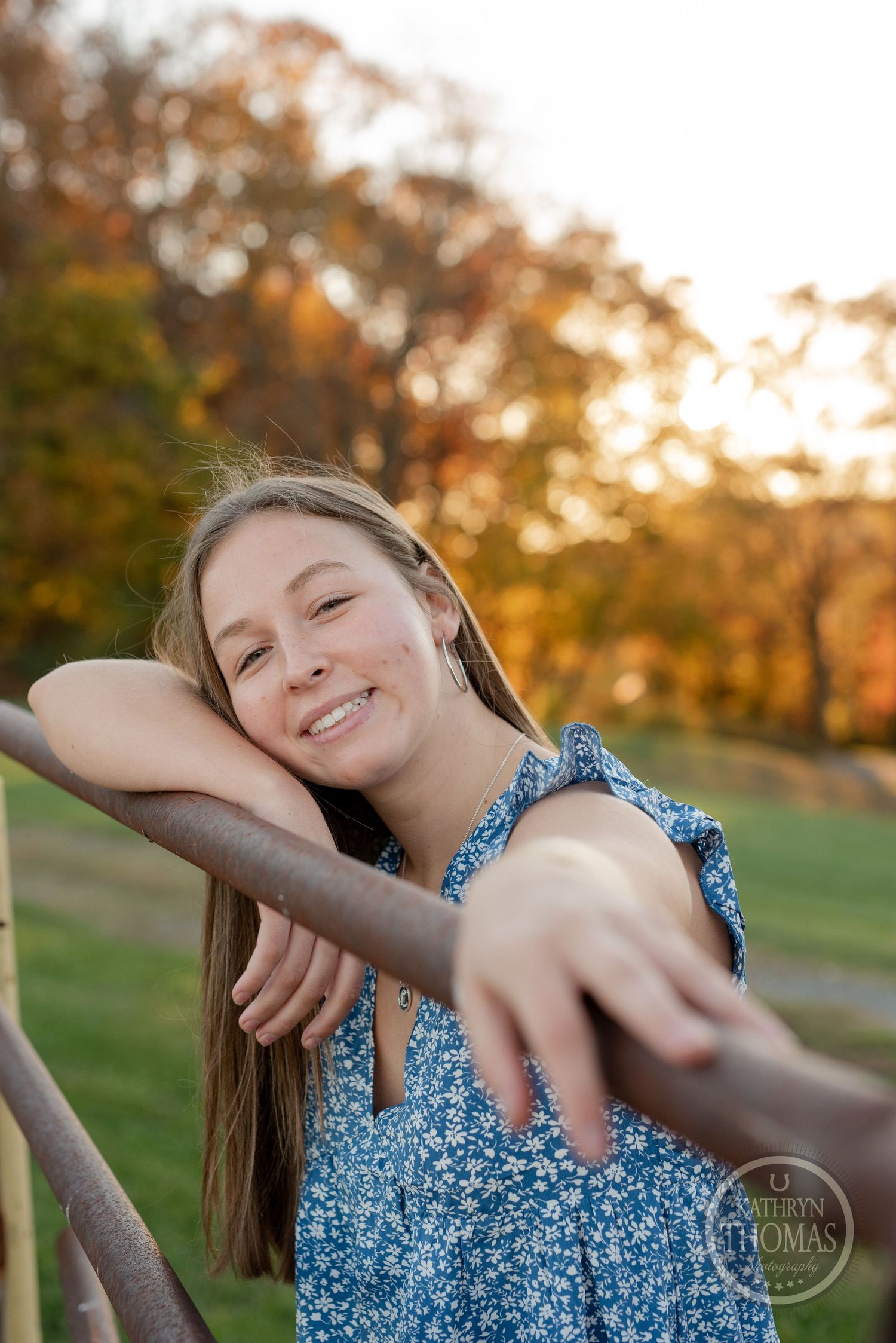 Someone in blue dress leans playfully on wooden railing with autumn trees behind.