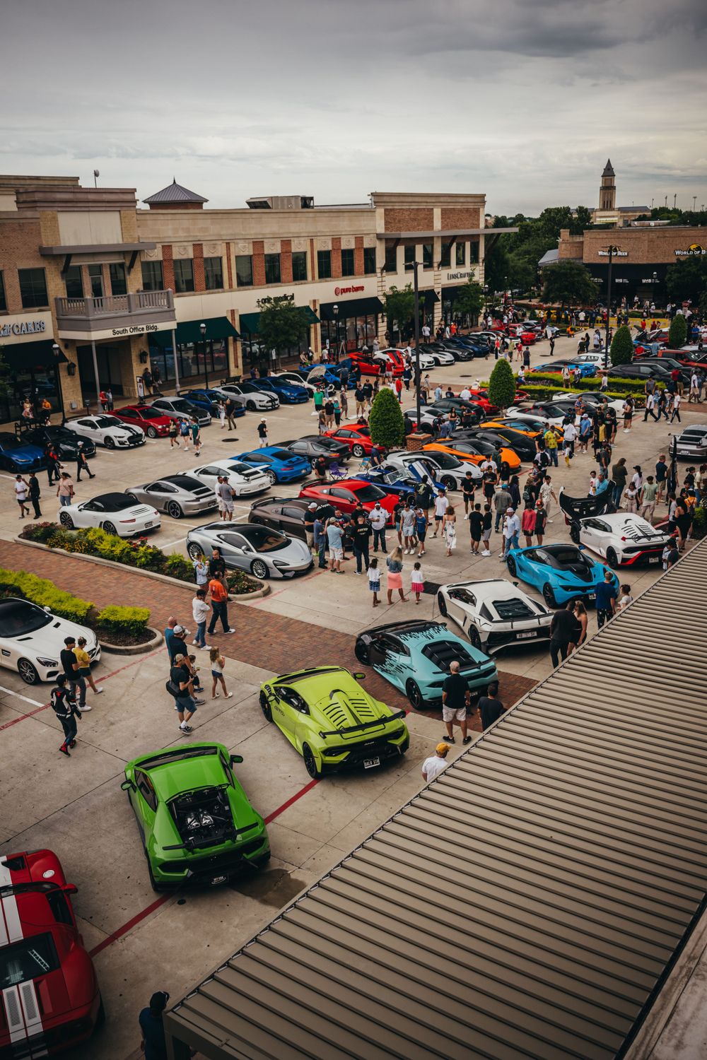 Aerial view of car show in shopping center parking lot with colorful vehicles on display.