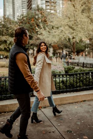 A couple walks through an urban park with city buildings visible in the background.