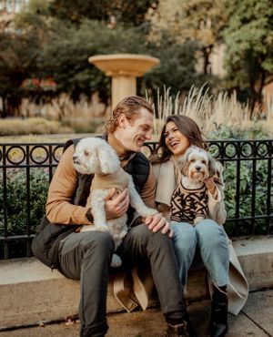 A couple laughs while sitting with their white dogs on a park bench during fall.