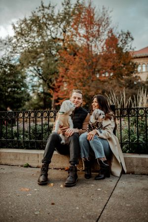 A couple sits on a stone wall with two small dogs on a beautiful fall day.