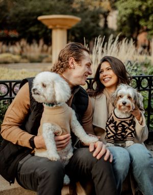 A couple smiles while sitting with their two small dogs on a stone wall during autumn.