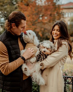 A couple holding their two small white dogs shares a loving moment in a park during autumn.