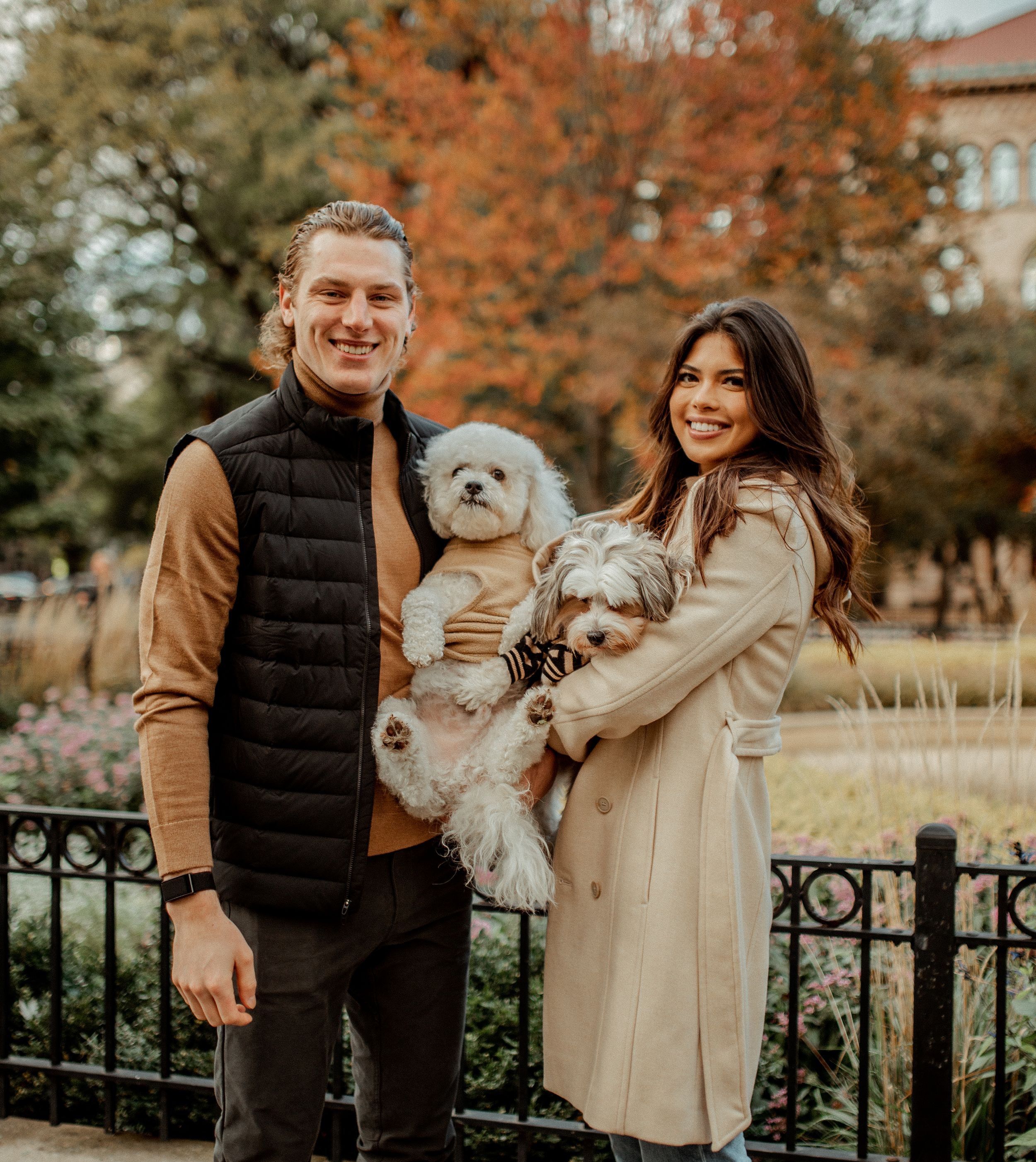 A couple poses with their two fluffy white dogs against a backdrop of fall colors and iron fencing.