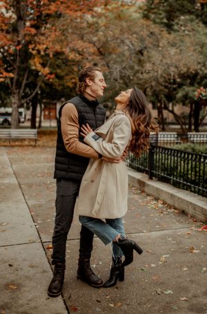 A couple shares a romantic moment on a sidewalk surrounded by fall trees.
