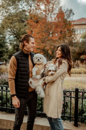 A couple smiles while holding their white puppies in a park surrounded by autumn trees.