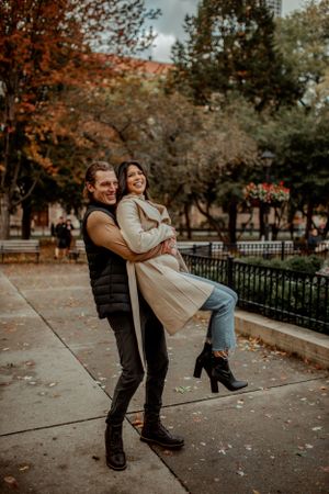 A playful couple embraces on a park pathway surrounded by autumn trees.