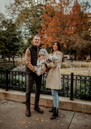 A couple poses with their small white dog on a fall day in an urban park setting.