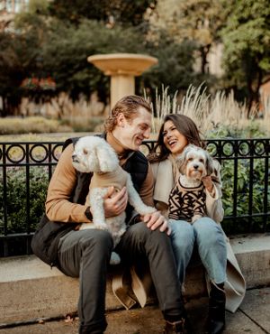 A couple sits on a park bench with two small white dogs, sharing laughs on an autumn day.