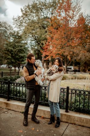 A couple in fall attire poses with their small white dogs in a park with autumn foliage.