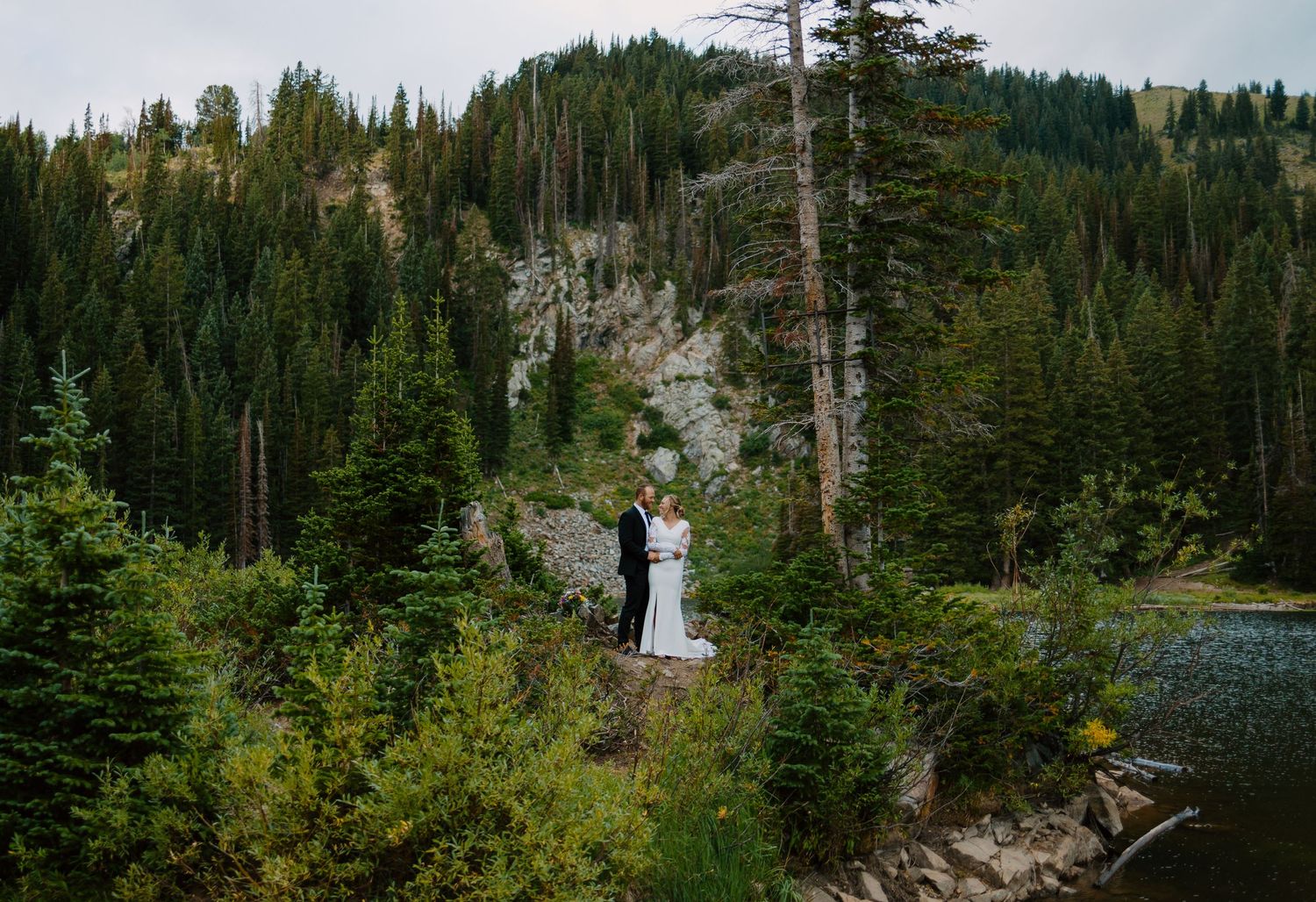 A couple stands together on a mountain ledge surrounded by evergreen forests and rocky cliffs.