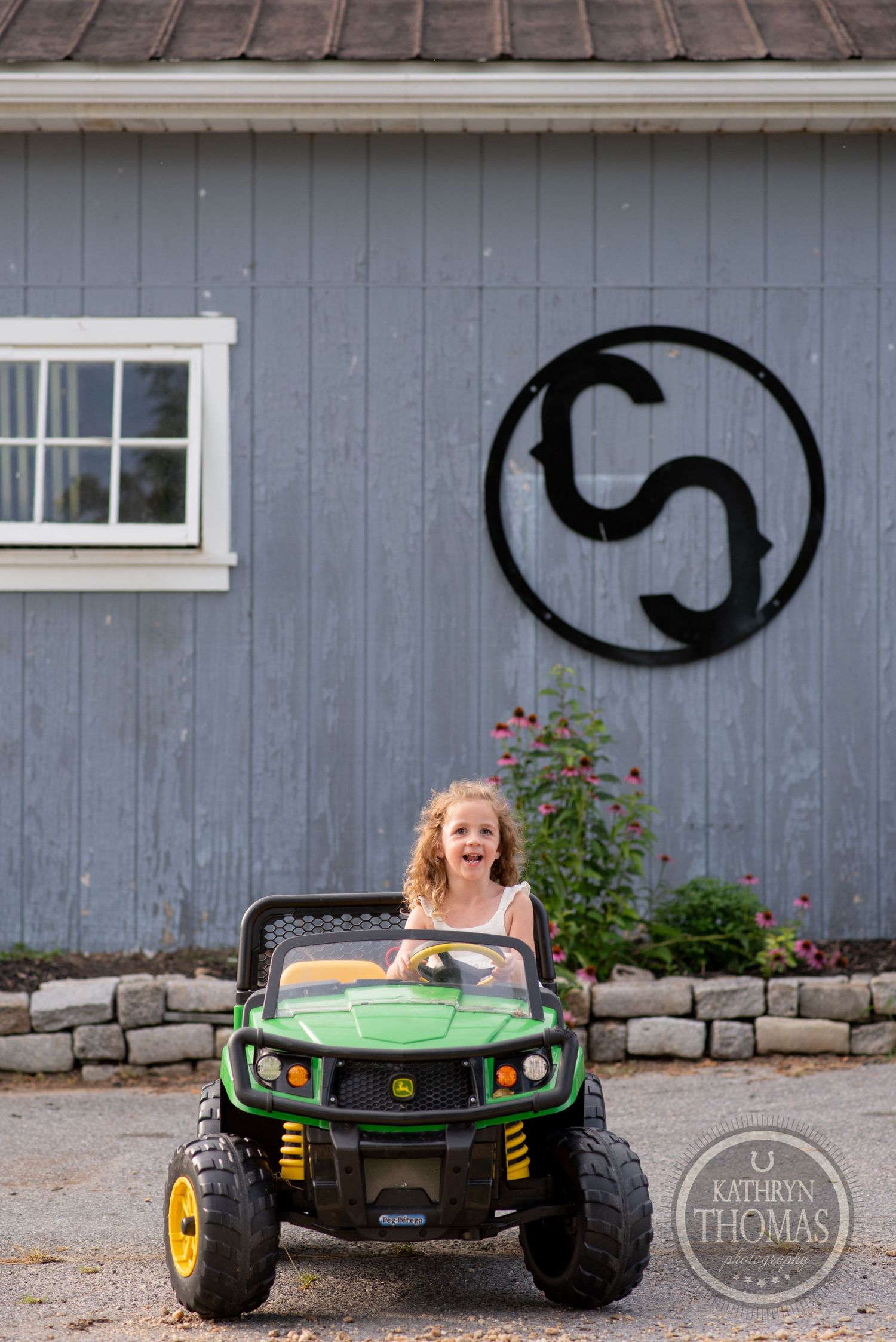 Child rides a green toy John Deere vehicle in front of a grey barn with circular logo.
