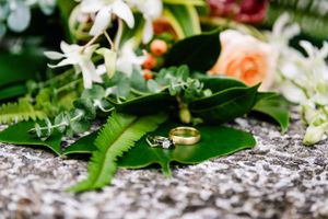 Wedding rings displayed with tropical leaves and flowers on a textured gray surface.