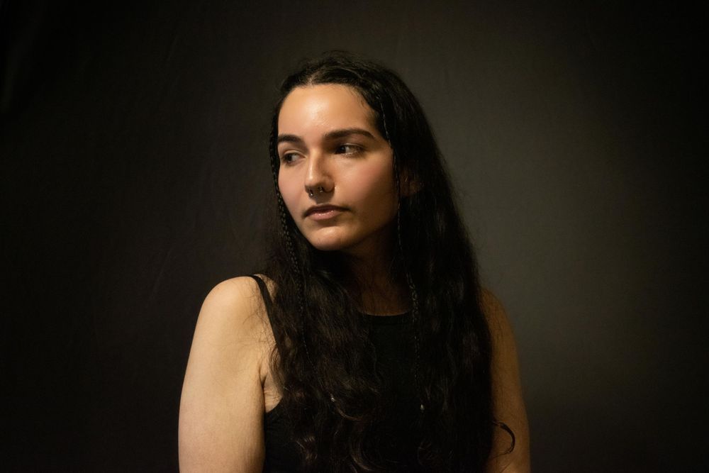 Moody side-lit portrait showing person with long dark hair against dark backdrop.