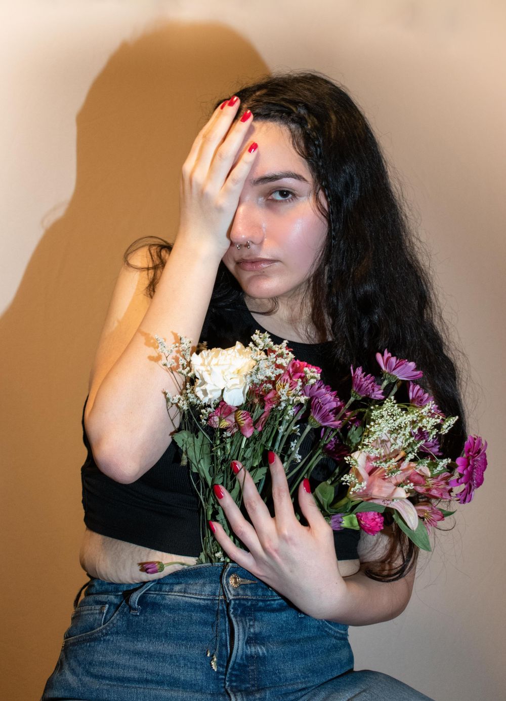 Artistic self-portrait with flowers and red nails against beige wall creating dramatic mood.