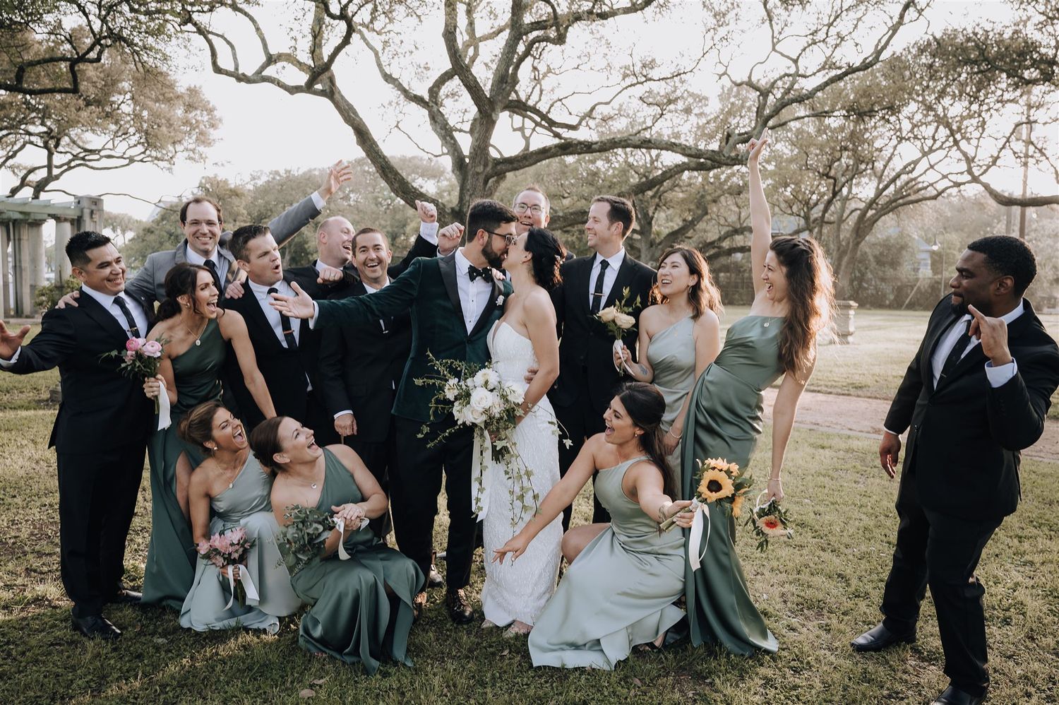 Large wedding party poses together on lawn during outdoor celebration.