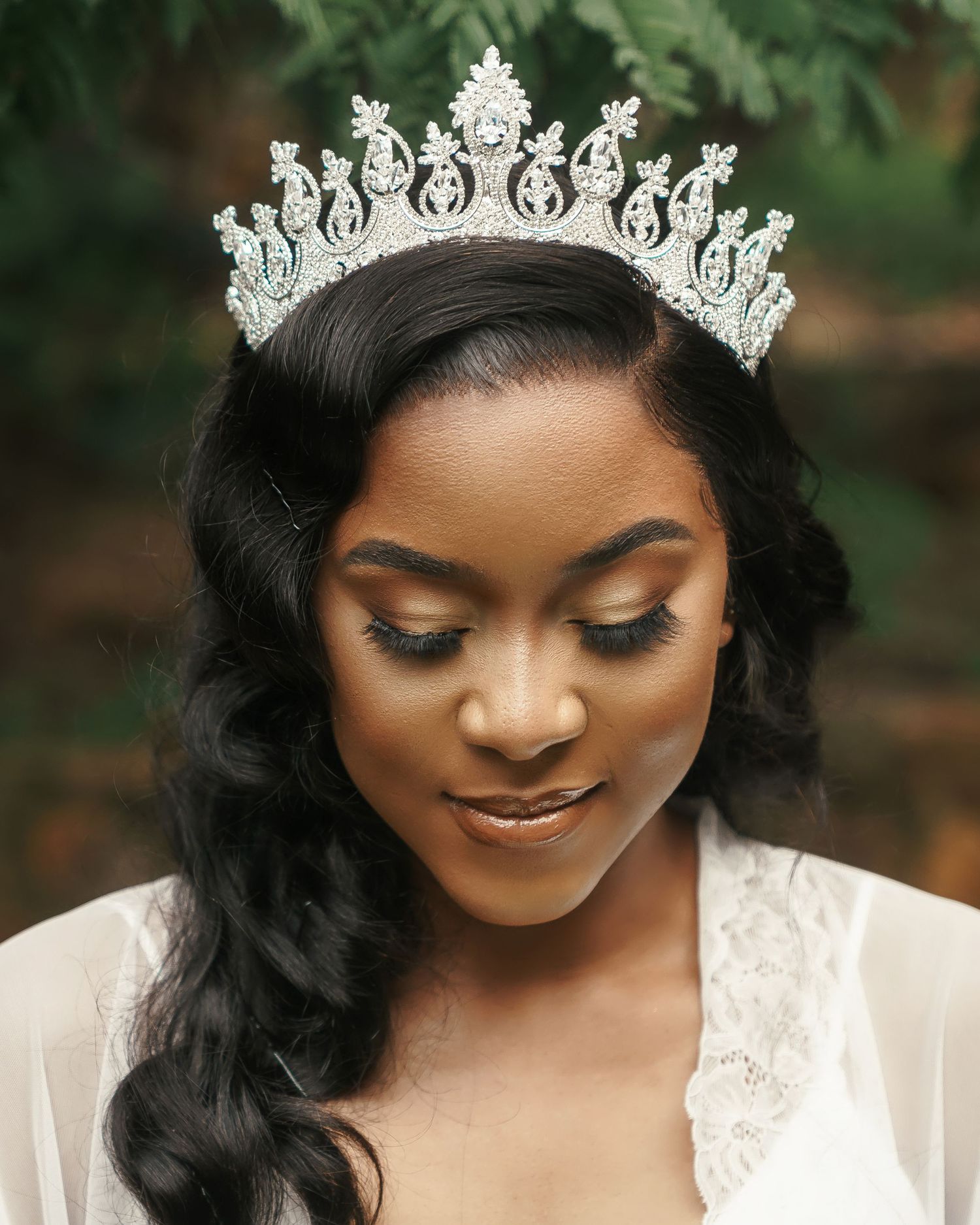 A jeweled tiara crown adorns dark flowing hair against a soft natural outdoor background.