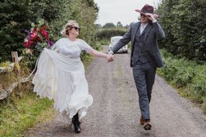 A couple in wedding dress and suit holding hands running toward the camera in front of Beetle bug car on a country path