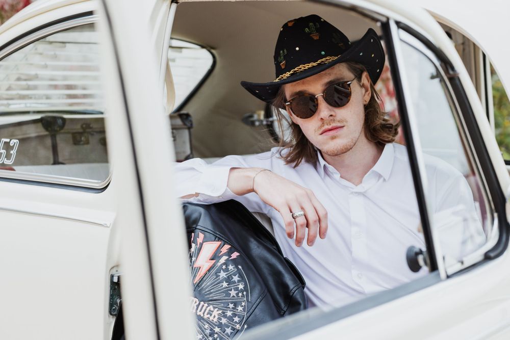 close up through the car window of Jack a man with blonde shoulder length hair in a cowboy hat and sunglasses