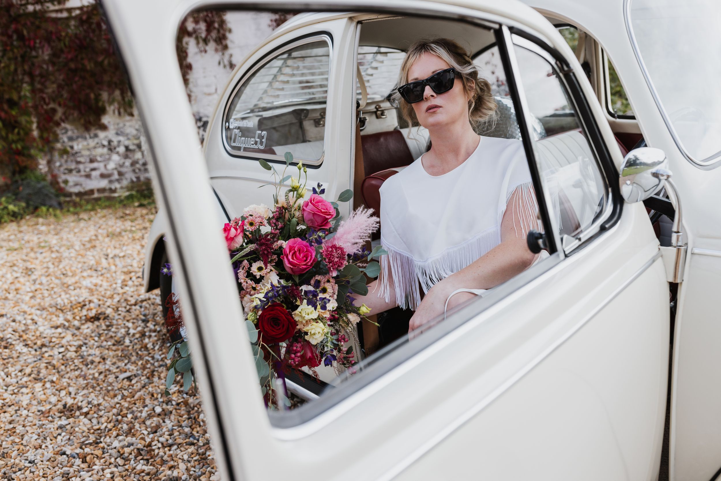 close up through the car window of female model with blonde hair in sunglasses holding a big bunch of colourful flowers.