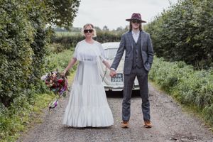 A couple in wedding dress and suit holding hands in front of Beetle bug car on a country path