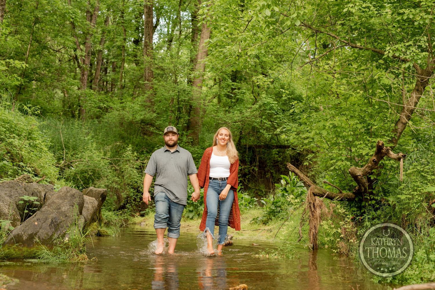 A couple wades barefoot in a creek surrounded by lush green forest scenery.