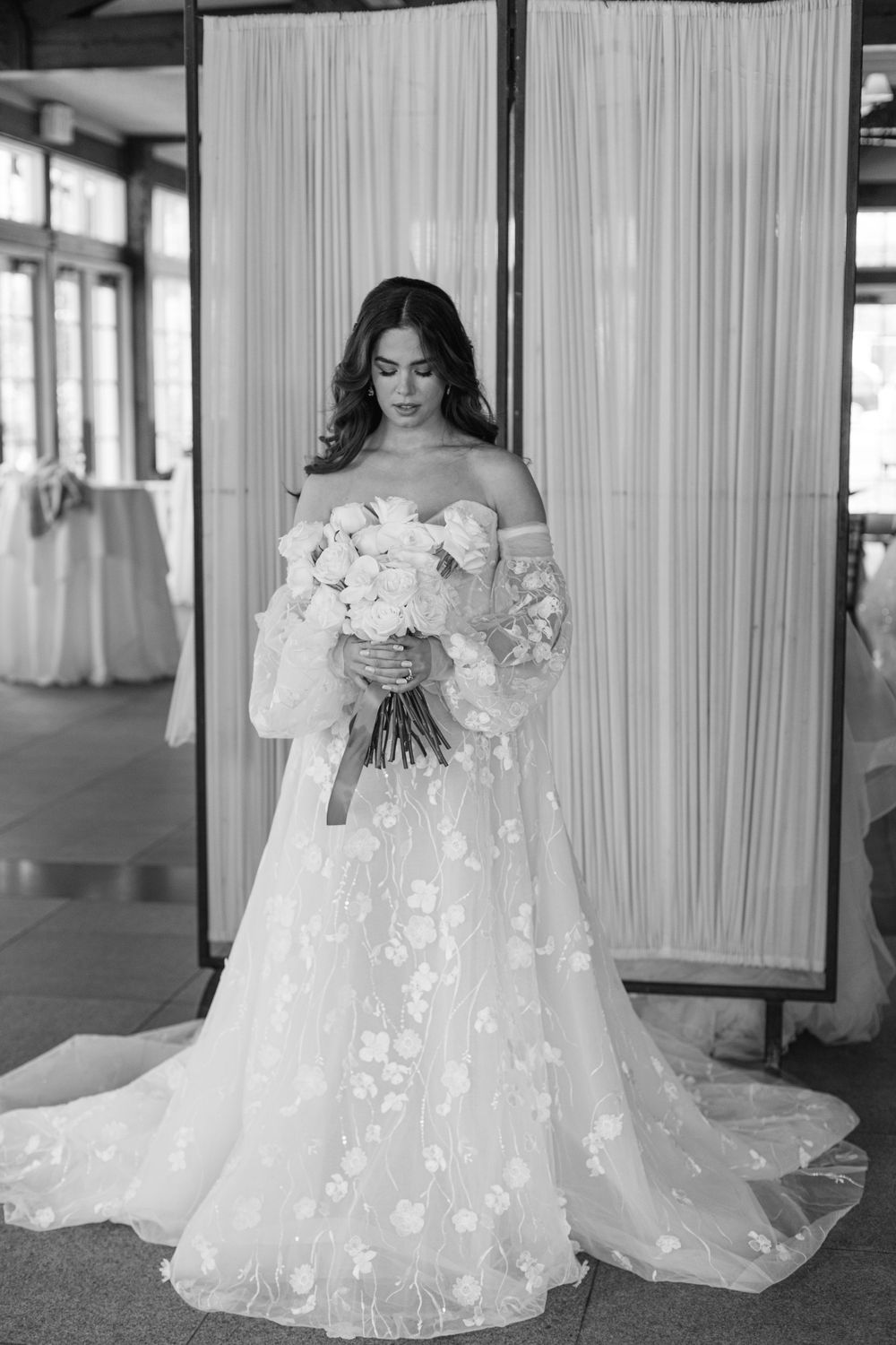 Bride in off-shoulder floral gown holds white bouquet against white curtain backdrop.