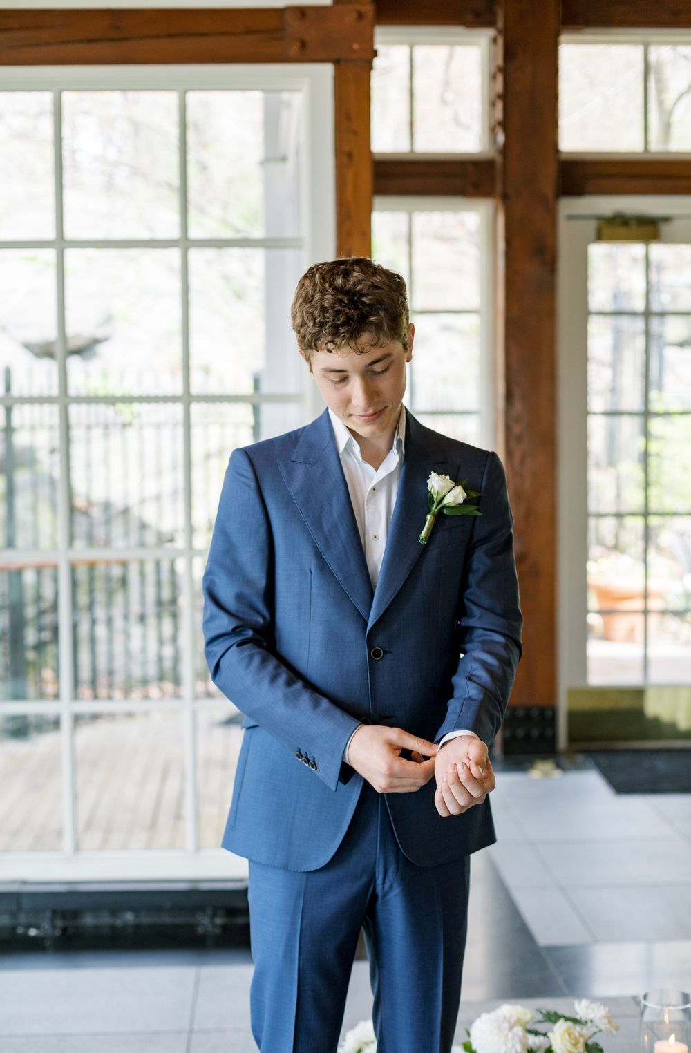 A well-dressed person in a blue suit adjusts their cuffs while standing in a sunlit room.