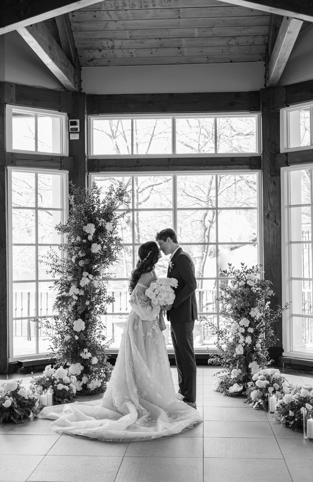 Wedding couple shares moment under floral arches by large windows in rustic venue.