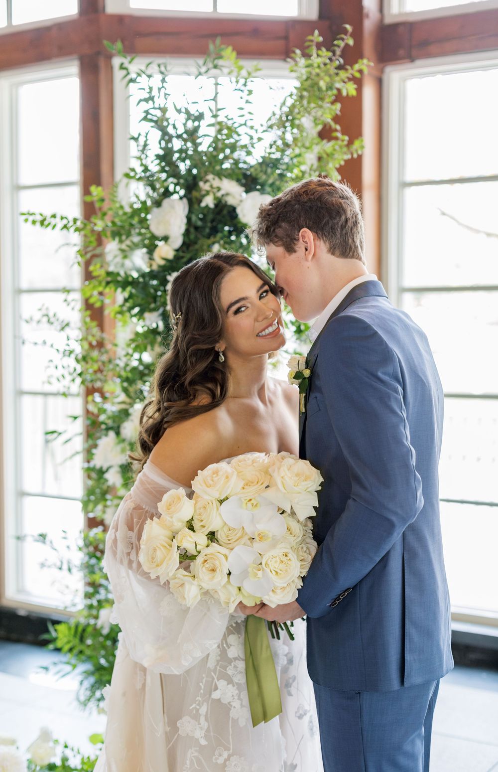 Joyful wedding couple poses with white bouquet by sunlit windows and greenery.